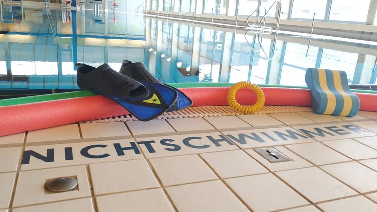 Swimming pool with fins, pool noodles, and a swimming board at the edge of the pool. On the tiles, it says 'NON-SWIMMER'. In the background, the water of the pool is visible.