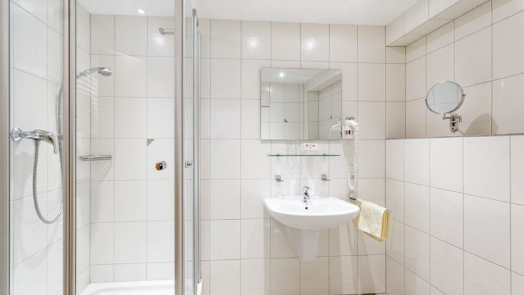Een modern badkamer met witte tegels en een douche. Er is een wastafel met spiegel en een handdoekhouder.