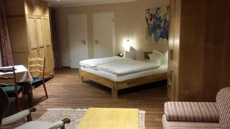 A cozy room with a double bed, a table, and chairs. The furnishings are wooden furniture, and there is a colorful piece of artwork on the wall.