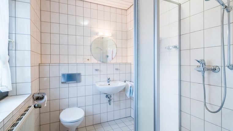 A modern bathroom with white tiles and a shower cabin. There is a sink, a toilet, and a large mirror.