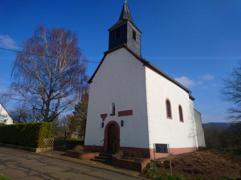 Eine kleine Kirche mit einem Turm und einem schwarzen Dach. Sie steht umgeben von Bäumen und blauem Himmel.