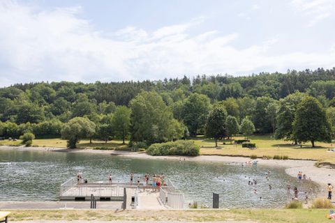 A tranquil lake surrounded by green trees and meadows. On the promenade, people can be seen relaxing and enjoying the water.