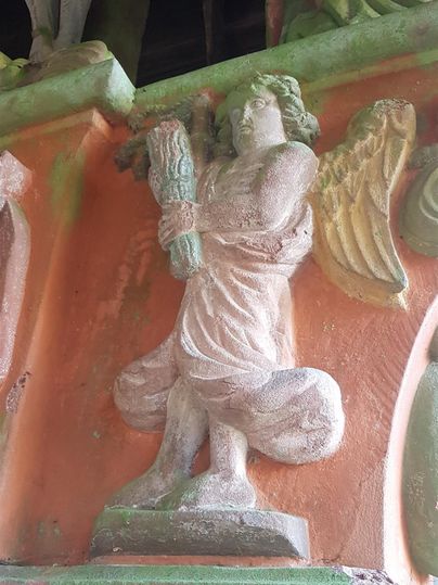 Eine Statue eines Engels mit Flügeln hält eine Blume. Der Hintergrund ist in warmen, erdigen Farben gehalten.