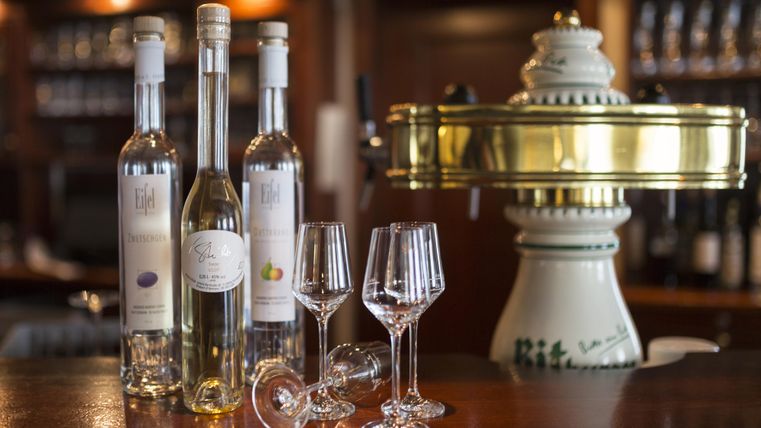 Auf der Theke stehen drei Flaschen Schnaps und zwei Gläser. Im Hintergrund ist eine elegante, nostalgische Spirituosenmaschine zu sehen.