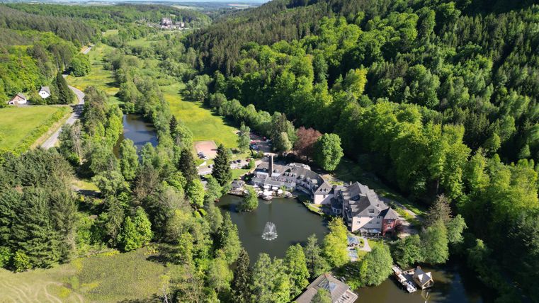 A picturesque landscape with a small lake surrounded by dense forests. In the background, gentle hills and a tranquil environment can be seen.