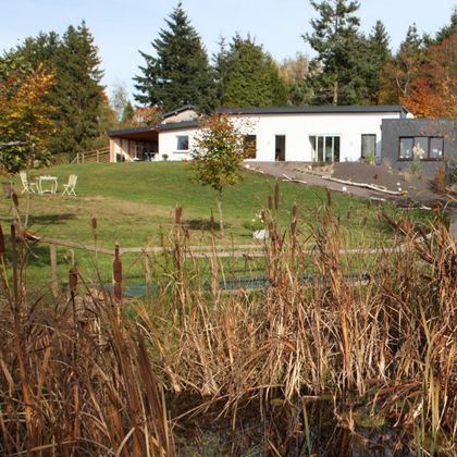 A modern house in the midst of a green landscape. In the foreground are tall plants and a seating area.