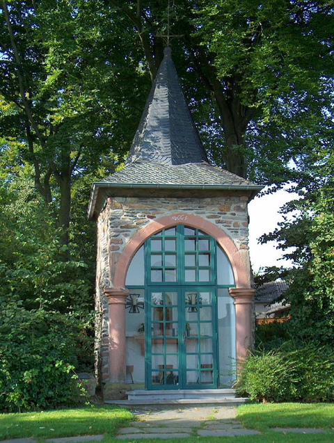 Ein historisches Gebäude mit einem spitzen Dach und großen Fenstern. Umgeben von grünen Bäumen und einem gepflegten Garten.