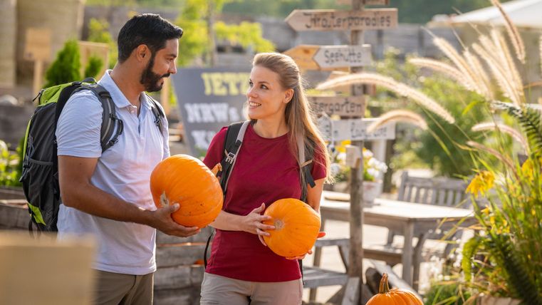 A couple is standing in a pumpkin field holding orange pumpkins. In the background, signs and garden furniture are visible.