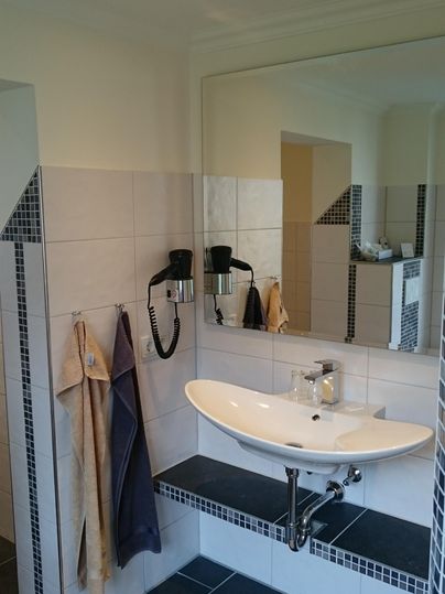 Ein modernes Badezimmer mit einem Waschbecken und großen Spiegeln. Die Wände sind gefliest und es gibt Handtücher an der Wand.