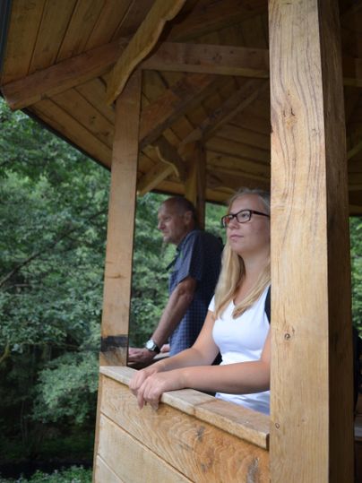 Ein Paar steht an einem Holz-Balkon und schaut in die Natur. Im Hintergrund sind Bäume zu sehen, die eine ruhige Atmosphäre schaffen.