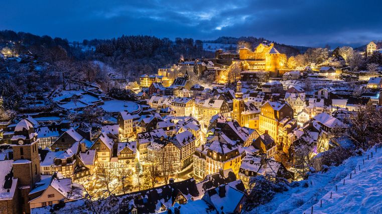 Eine malerische Stadt im Winter, überzogen mit Schnee und beleuchtet von warmem Licht. Im Hintergrund sieht man eine alte Burg, umgeben von schneebedeckten Bäumen.