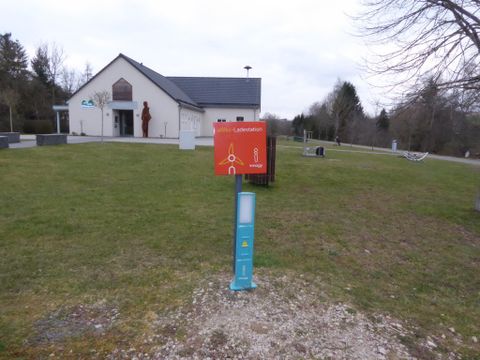 Ein Gebäude mit einem grauen Dach steht auf einer grünen Wiese. Vor dem Gebäude befindet sich ein Informationsschild.