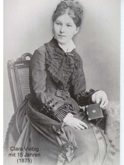 Ein sepiafarbenes Porträt eines jungen Mädchens, das im Jahr 1875 aufgenommen wurde. Sie trägt ein elegantes, viktorianisches Kleid und hält ein kleines Fotobuch in der Hand.