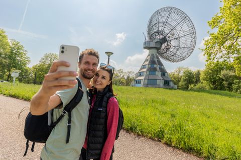 Ein glückliches Paar macht ein Selfie vor einer großen Satellitenschüssel. Im Hintergrund sind grünes Gras und Bäume zu sehen.