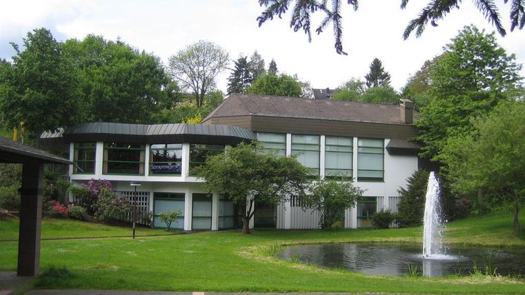 Een modern gebouw omgeven door een groene landschapstuin. Op de voorgrond is een kleine vijver met een waterfontein.