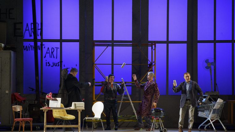 Een podiumvoorstelling met meerdere acteurs die verschillende rekwisieten vasthouden. De achtergrond is in levendig blauw vormgegeven.