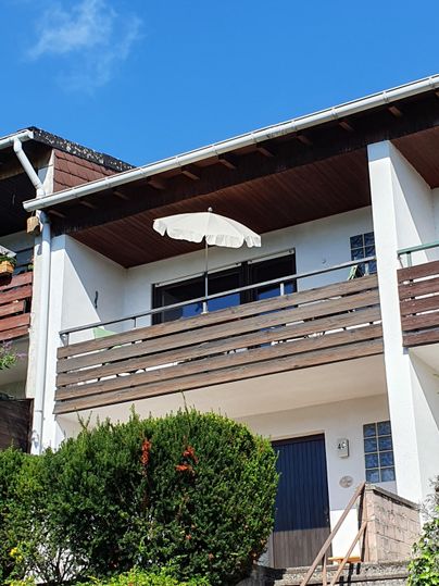 Ein modernes Haus mit einem Balkon und einem Sonnenschirm. Die Umgebung ist grün mit Bäumen und Sträuchern.