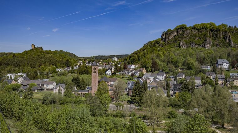 Panoramablick auf eine Stadt mit einer Kirche im Vordergrund und bewaldeten Felsen im Hintergrund unter blauem Himmel.
