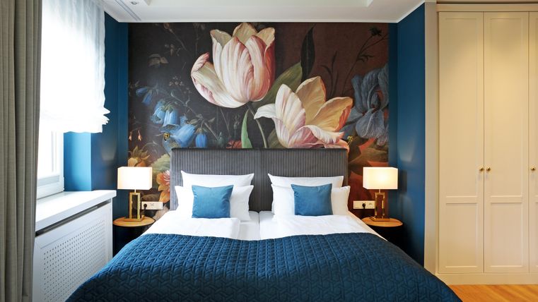 Ein modernes Schlafzimmer mit einem großen Bett und bunten Blumenbildern an der Wand. Die Einrichtung ist elegant und minimalistisch mit warmen Holz- und Blautönen.