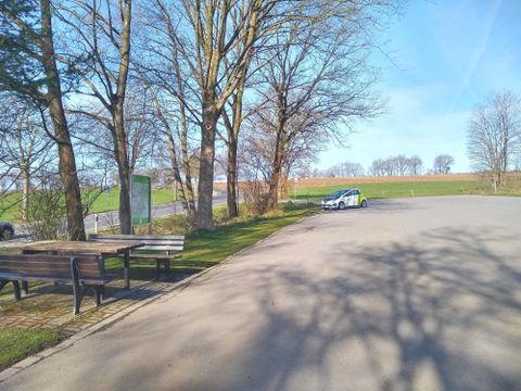Een rustige parkeerplaats omringd door bomen en weilanden. De zon schijnt en de lucht is helder.