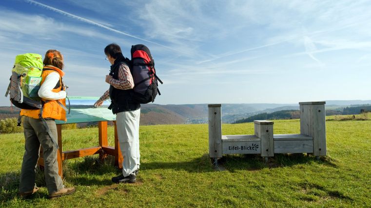 Zwei Wanderer mit Rucksäcken betrachten eine Informationstafel in einer hügeligen Landschaft mit weitem Blick.
