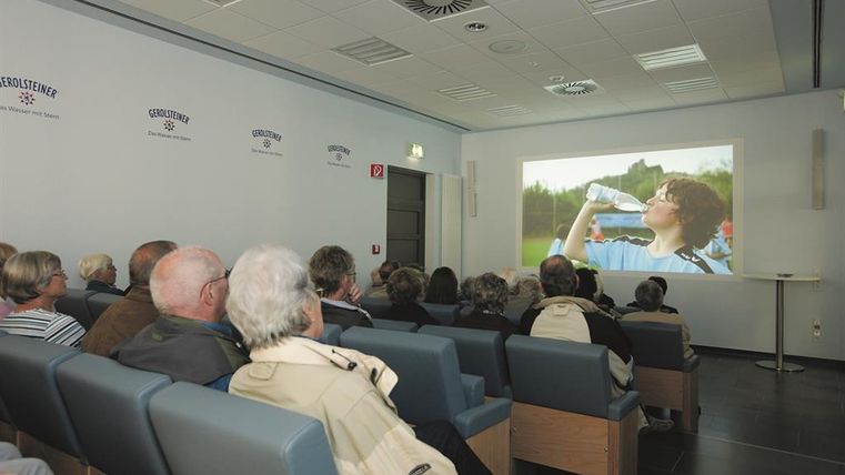 People in a small cinema are watching a film about Gerolsteiner water. Gerolsteiner logos are visible on the wall.