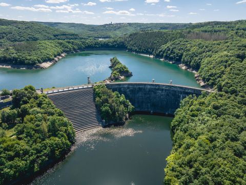 A dam surrounded by lush greenery and a calm lake. The sky is clear and blue, creating a peaceful atmosphere.