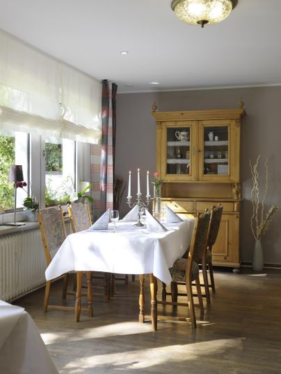 Ein gemütlicher Essbereich mit einem Tisch, der mit einer weißen Tischdecke gedeckt ist. Im Hintergrund steht ein Holzwandschrank und große Fenster lassen viel Licht herein.