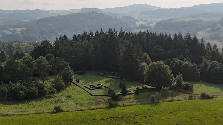 Luftaufnahme eines Waldfriedhofs mit geometrischen Strukturen, umgeben von Bäumen und Wiesen.
