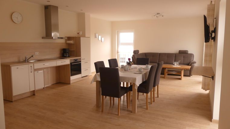 Eine moderne, hell erleuchtete Küche mit angrenzendem Essbereich. Ein großer Tisch mit Stühlen steht in der Mitte, und im Hintergrund ist eine gemütliche Sitzecke.