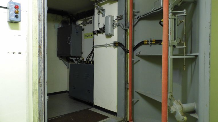 Ein Raum mit technischen Geräten und einem offenen Zugang. An den Wänden sind Rohre und Kabel sichtbar.