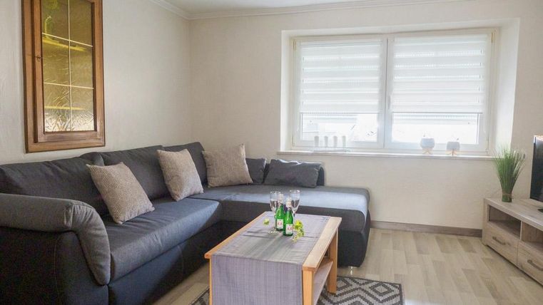 Ein gemütliches Wohnzimmer mit einer grauen Couch und einem Holzkaffee-Tisch. Das Fenster lässt viel Licht herein und es gibt eine Pflanze in der Ecke.