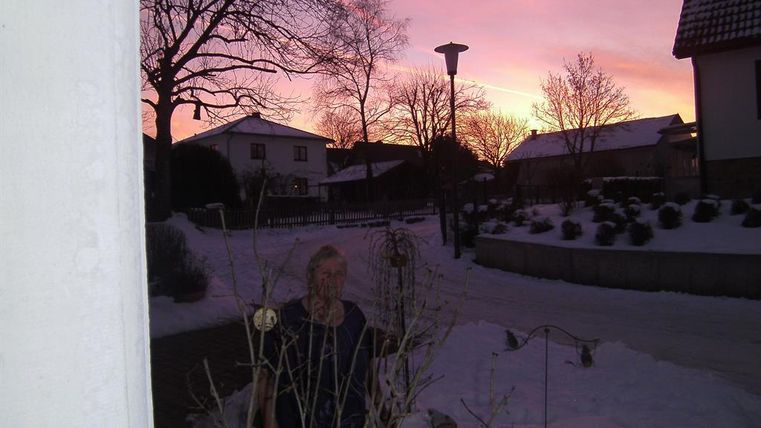 A snow-covered street with houses and trees in the background. The sky is painted in soft shades of pink and violet.