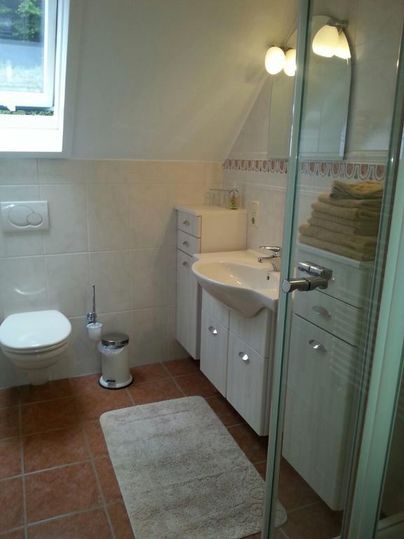 Ein modernes Badezimmer mit einem Waschbecken, einer Dusche und weißen Möbeln. Braune Fliesen und ein großes Fenster sorgen für ein helles Ambiente.