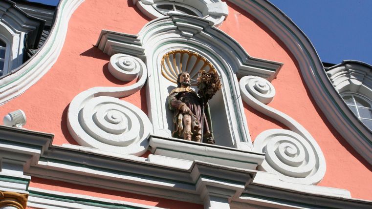 Eine dekorative Fassade mit einer farbigen Wand und kunstvollen Verzierungen. In der Mitte befindet sich eine Statue, die einen historisch gekleideten Mann darstellt.