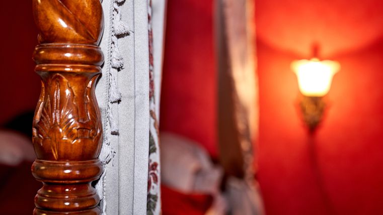 A detailed wooden post of a bed stands in the foreground. In the background, one can see a warm light source on a red-painted wall.