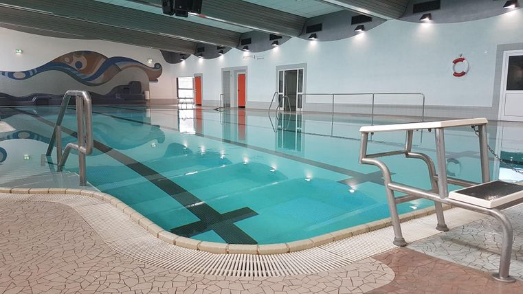 A modern indoor swimming pool with clear water and a wide staircase. The walls are designed in light colors and there is safety equipment nearby.