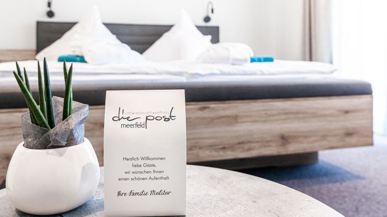 Ein gemütliches Schlafzimmer mit einem großen Bett und hellen Laken. Auf dem Tisch steht eine Willkommenskarte und eine Pflanze in einem Topf.