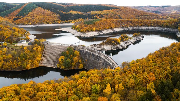 Eine beeindruckende Landschaft mit einem Stausee und bunten Wäldern im Herbst. Die Dammmauer erstreckt sich über das ruhige Wasser.