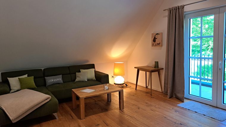 A cozy living room with a green couch and a wooden table. Large windows let in plenty of light and offer a view outside.
