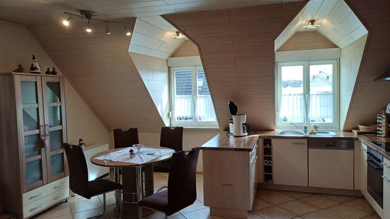 A modern kitchen with sloped walls and large windows. There is a table with chairs and a kitchenette with appliances.