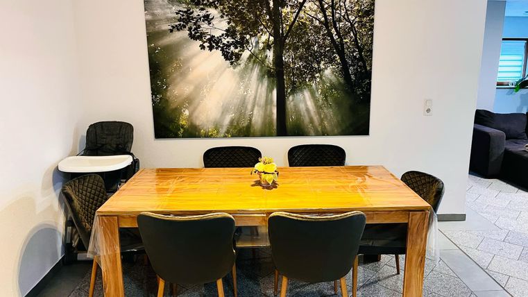 A cozy dining area with a wooden table and black chairs. A large picture of trees with sun rays hangs on the wall.