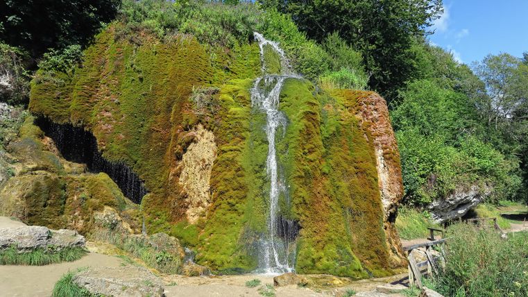 Een kleine waterval stroomt over een met mos bedekte rotsformatie. Omgeven door weelderige, groene plantengroei en een blauwe lucht.