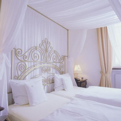 An elegant bedroom with two single beds and an ornate headboard. The room is bright and cheerful, with soft curtains and warm lighting.