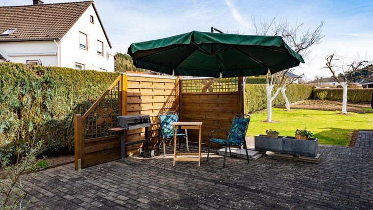 Ein gemütlicher Gartenbereich mit einem Tisch, zwei Stühlen und einem Sonnenschirm. Es gibt eine gepflasterte Fläche und einen grünen Rasen im Hintergrund.