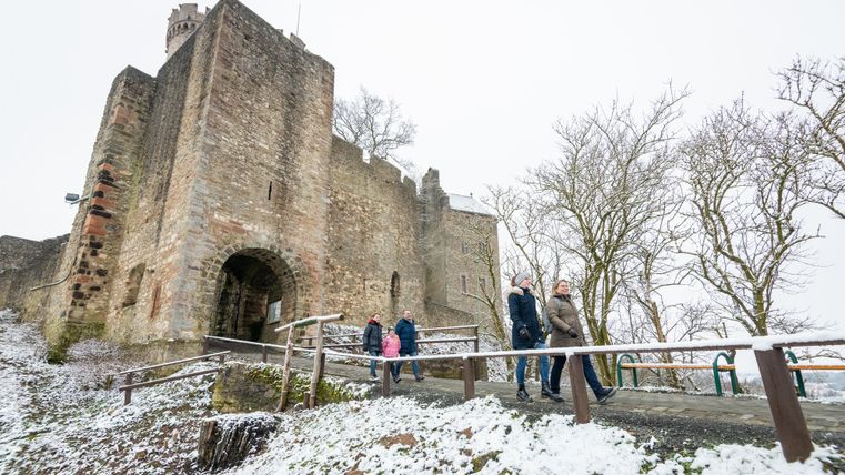 Eine alte Burg umgeben von Schnee und kahlen Bäumen. Menschen gehen den Weg zur Burg hinauf.