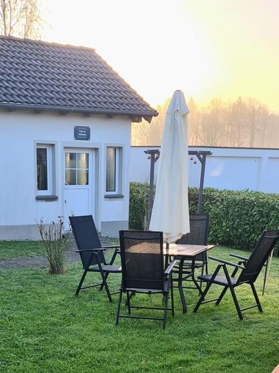 Ein gemütlicher Außenbereich mit einem Tisch und Stühlen. Im Hintergrund steht ein kleines Gebäude, umgeben von grünem Rasen und sanftem Licht.