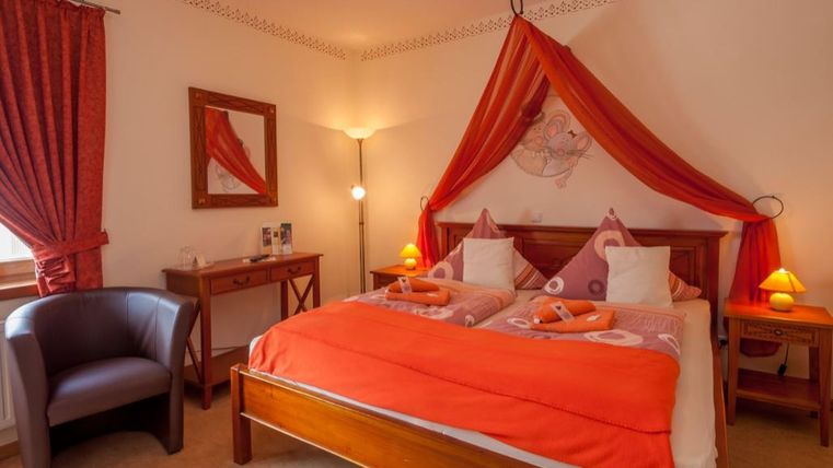 Ein gemütliches Schlafzimmer mit einem großen Bett in Orange- und Weißtönen. Die Einrichtung umfasst einen Sessel und dekorative Vorhänge, die eine warme Atmosphäre schaffen.
