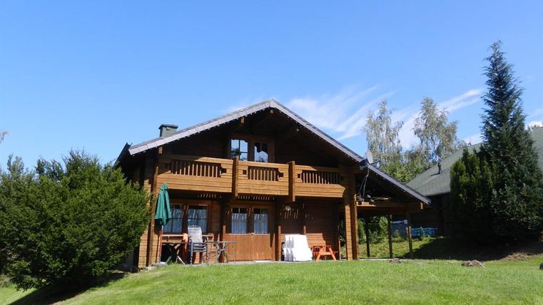 Ein gemütliches Holzhaus mit Balkon, umgeben von grünem Rasen und Bäumen. Der Himmel ist blau und klar.