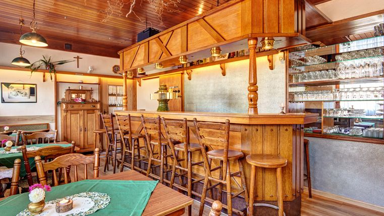 Ein gemütliches Restaurant mit einem Holzbar und leeren Stühlen. Die Tische sind mit grünen Tischdecken gedeckt und es gibt eine sichtbare Auswahl an Gläsern im Hintergrund.